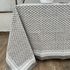 May include: A rectangular tablecloth featuring a grey background with a repeating white floral block print pattern. The fabric has a wide solid grey border with a thin white geometric trim. A white bowl with colorful floral accents holds red apples.