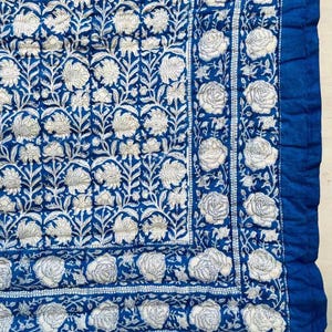 May include: A quilted textile featuring a deep blue background with a repeating white floral pattern. The central area displays dense botanical motifs, while the border showcases a series of individual rose-like blossoms in a linear arrangement.