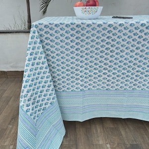 May include: A white tablecloth with a repeating blue floral pattern. The tablecloth has a border with a geometric design in shades of blue and green. A bowl of red apples sits on the table.
