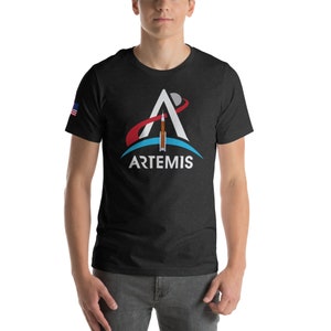 Op de afbeelding: Een zwart T-shirt met een witte afbeelding van het Artemis-programma logo. Het logo toont een witte letter A met een rode boog en een blauwe cirkel eromheen. Een raket bevindt zich in de A. De tekst "ARTEMIS" staat onder het logo.