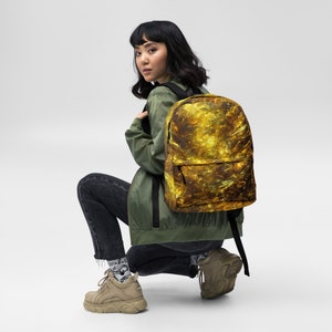 May include: A person wearing a green jacket and black jeans is wearing a backpack with a gold and black abstract pattern. The backpack has a zipped pocket on the front.