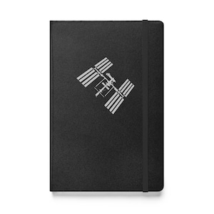 May include: Black notebook with a silver graphic of the International Space Station. The notebook has a black elastic band closure and a textured cover. The design is a simple, modern style.