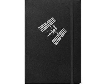 May include: Black notebook featuring a silver graphic of the International Space Station. The notebook has a black elastic closure and a textured cover. The design is a simple, modern style.