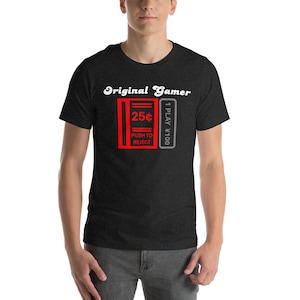May include: A dark heather grey t-shirt with a white graphic of a retro arcade game machine. The graphic says "Original Gamer" and "25¢ Push to Reject" and "1 Play $100".