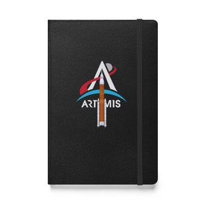 Op de afbeelding: Een zwart lederen notitieboek met een wit en blauw Artemis-logo met een raket en de tekst "ARTEMIS".