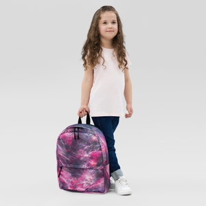 Puede incluir: Una mochila de galaxia rosa y morada con correas y tiradores de cremallera negros. Una persona joven sostiene la mochila.