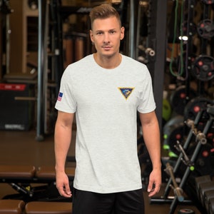 May include: A heather grey t-shirt with a blue and yellow triangle patch featuring a helicopter and the text "The Brotherhood of the Winged Wheel". The t-shirt also has a small American flag patch on the left sleeve.