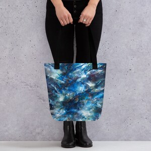 May include: A tote bag with a blue, white, and green abstract design. The bag has black straps and is being held by a person.