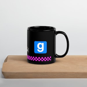 May include: Black ceramic mug featuring a blue square with a white "g" and a pink and black checkered pattern. The mug is displayed on a wooden surface.