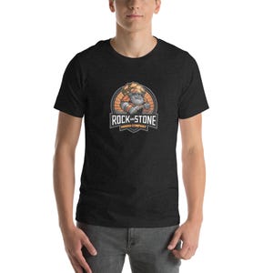 May include: Dark gray t-shirt featuring a circular logo with a cartoon miner holding a pickaxe. The logo includes the text "ROCK AND STONE MINING COMPANY" in white and orange. The shirt is short-sleeved and has a crew neck.