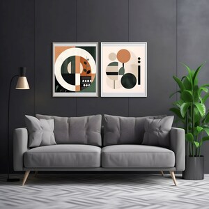 Puede incluir: Dos impresiones de arte geométrico abstracto con fondo negro, formas verdes, naranjas y blancas. Las impresiones están enmarcadas en un marco de madera claro y están colgadas en una pared gris oscuro sobre un sofá gris.