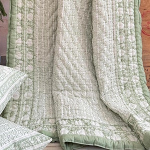 Könnte beinhalten: Eine grün-weiße gesteppte Decke mit floralem Muster. Die Decke ist gefaltet und über ein Kissen gelegt.