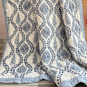 Puede incluir: Una colcha acolchada blanca y azul con un patrón repetido de árboles estilizados y motivos florales. La colcha tiene un borde azul claro y un patrón de costura en forma de diamante. El diseño es una mezcla de azul y blanco, creando un aspecto clásico y elegante.