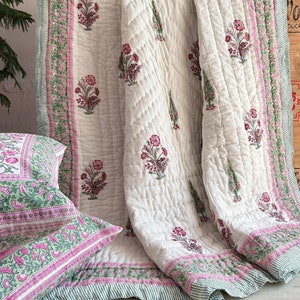 Op de afbeelding: Een wit katoenen dekbed met een bloemenprint in roze en groene tinten. Het dekbed heeft een gestikt patroon en een rand met een gestreept ontwerp. Het dekbed is gevouwen en wordt op een oppervlak getoond.