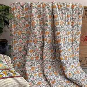 May include: A white cotton quilt with a floral print in shades of pink, yellow, and green. The quilt is folded and draped over a wooden surface.