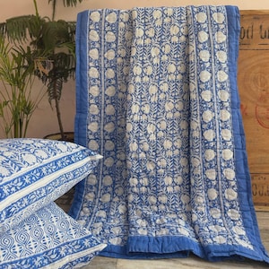 May include: A blue and white floral patterned quilt and two matching throw pillows. The quilt has a blue border and a repeating white floral design. The pillows have a similar pattern. The bedding is likely cotton.