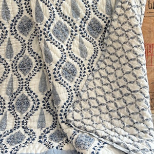May include: A white and blue quilt with a repeating pattern of stylized trees and floral motifs. The quilt features a light blue border and a quilted design. The reverse side has a geometric pattern in the same color scheme. This bedding item is suitable for a bed or as a decorative throw.