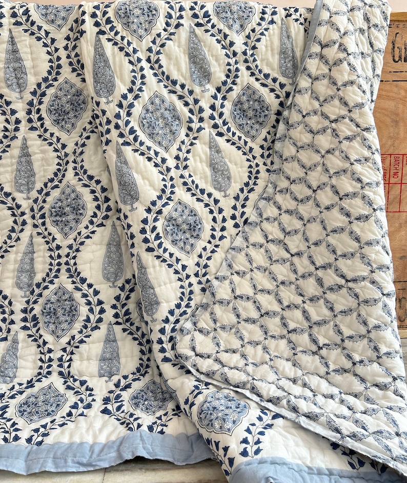 May include: A white and blue quilt with a repeating pattern of stylized trees and floral motifs. The quilt features a light blue border and a quilted design. The reverse side has a geometric pattern in the same color scheme. This bedding item is suitable for a bed or as a decorative throw.