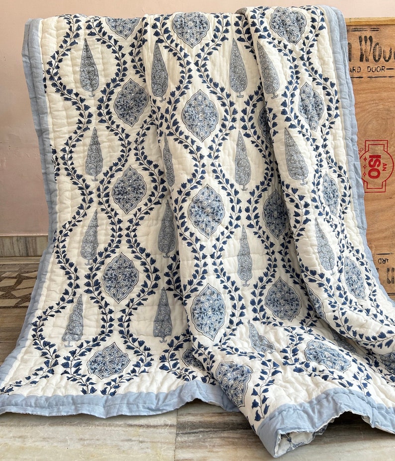 May include: A white and blue quilted throw blanket with a repeating pattern of stylized trees and floral motifs. The blanket has a light blue border and a diamond-stitched pattern. The design is a mix of blue and white, creating a classic, elegant look.