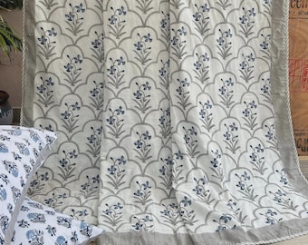 Könnte beinhalten: Ein weißes Steppbett mit passendem Kissenbezug, das ein sich wiederholendes Muster aus blauen Blumenzweigen in geschwungenen grauen Umrandungen zeigt. Die Bettwäsche hat einen einfarbigen grauen Rand mit einer dünnen weiß gestreiften Kante.