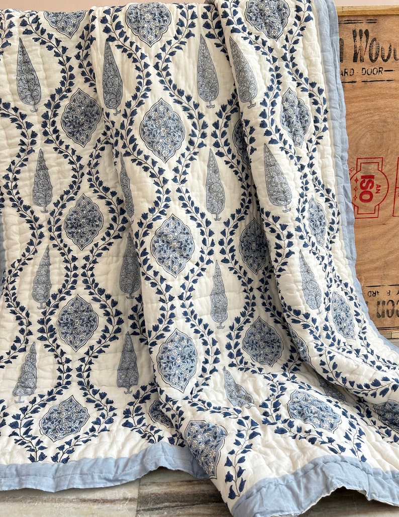 May include: A white and blue patterned quilt with a repeating design of stylized floral and tree motifs. The quilt features a light blue border and is made of a soft, quilted fabric. The overall design is symmetrical and detailed.