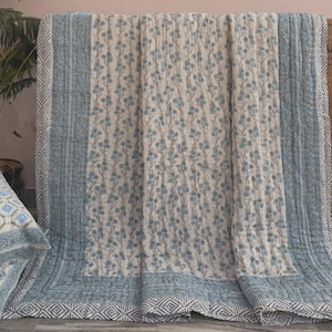 May include: A cream-colored quilt with a blue floral pattern and a blue border. The border features a geometric diamond design. The quilt is draped over a surface, showcasing its texture and design.