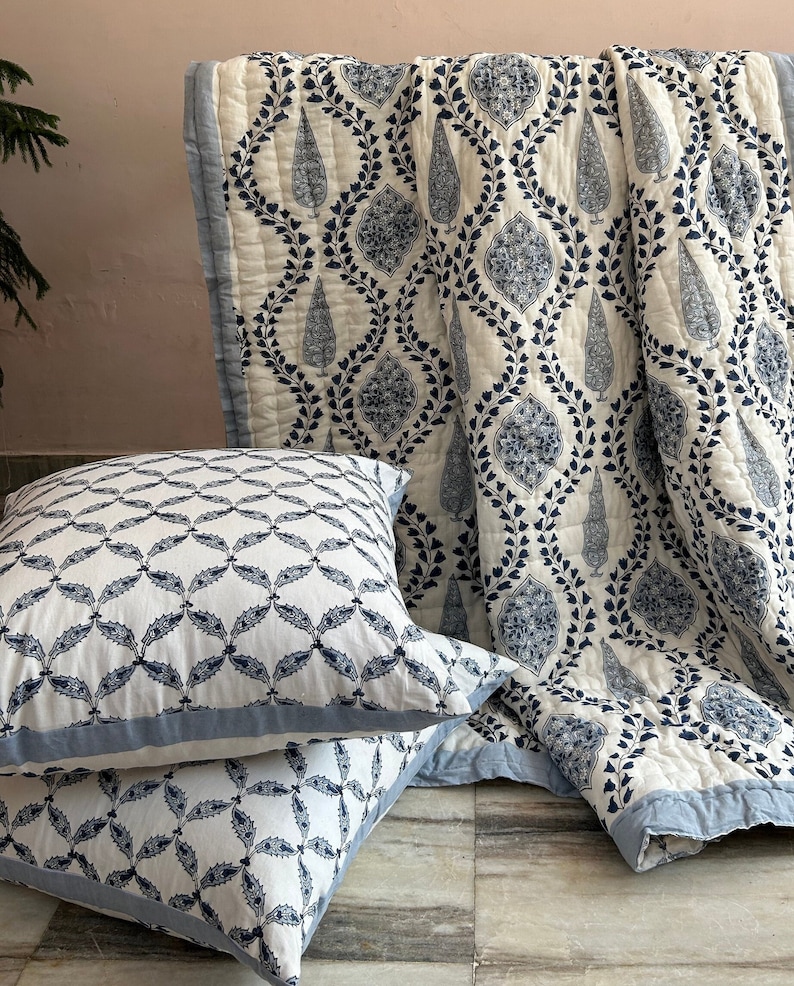 May include: A white and blue patterned quilt and two matching throw pillows. The quilt features a repeating design of teardrop shapes and floral elements in navy blue. The pillows have a similar pattern with a blue border. The bedding is arranged on a light-colored surface.