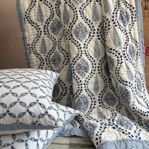 May include: A white and blue patterned quilt and two matching pillows. The quilt features a repeating design of stylized leaves and floral motifs in navy blue. The pillows have a similar pattern, with a blue border. The bedding set is made of a soft, quilted fabric.