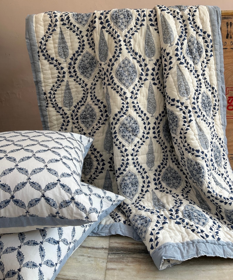May include: A white and blue patterned quilt and two matching pillows. The quilt features a repeating design of stylized leaves and floral motifs in navy blue. The pillows have a similar pattern, with a blue border. The bedding set is made of a soft, quilted fabric.