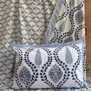May include: A white pillow with a blue floral design and a light blue border. The pillow is part of a bedding set with matching patterned fabric in the background. The pattern includes teardrop and floral motifs.
