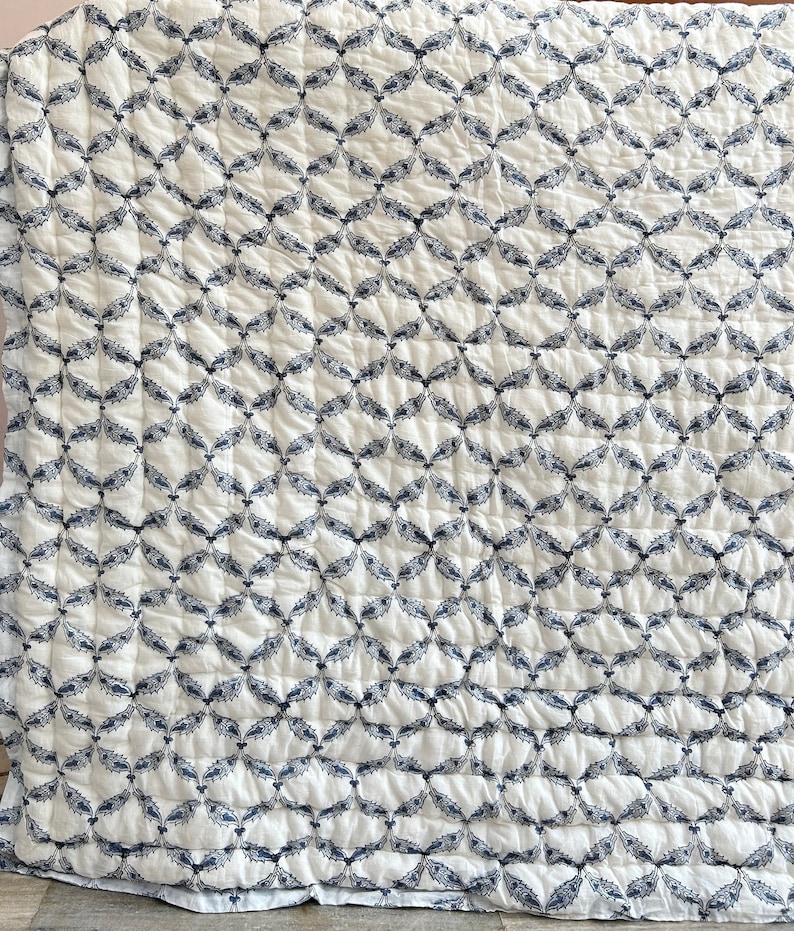 May include: White quilt with a repeating pattern of navy blue leaf-like shapes. The quilt has a quilted texture, with visible stitching creating a grid pattern across the surface. The overall design is symmetrical and visually appealing.