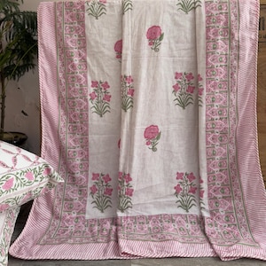 以下が含まれることがあります： ピンクのバラと緑の葉のデザインが施された白い花柄のベッドスプレッド。ベッドスプレッドにはピンクと白のストライプのボーダーと装飾的な花のボーダーがあります。お揃いの枕が隅に見えます。