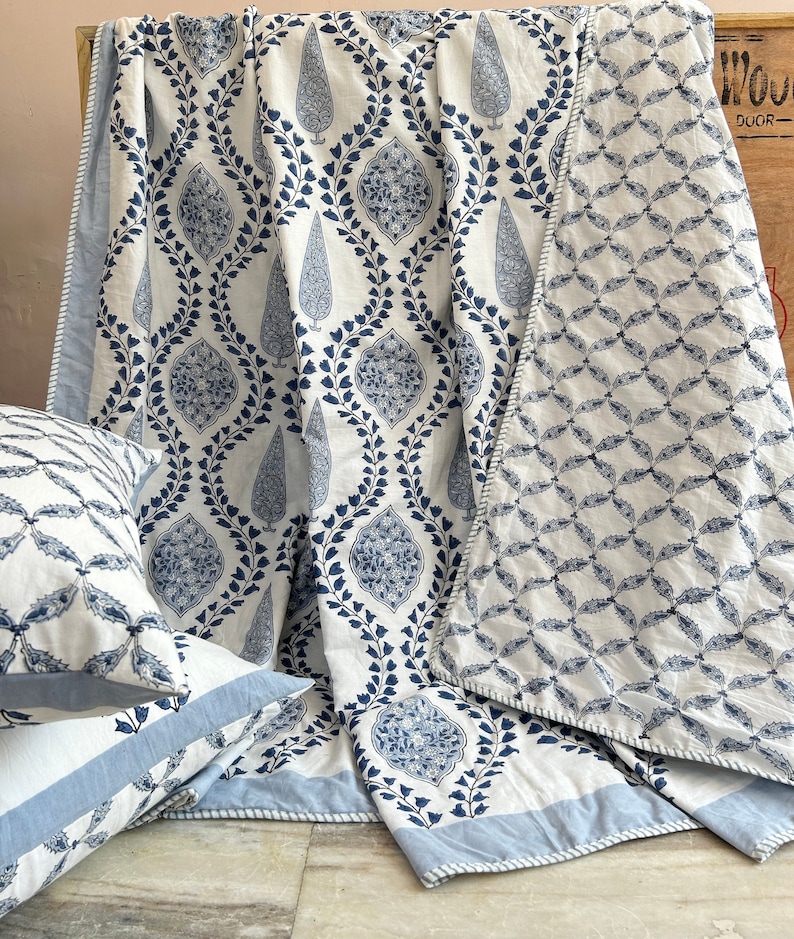 May include: A white and blue patterned quilt and matching pillow set. The quilt features a repeating design of blue floral motifs on a white background. The pillows have a similar pattern. The set is made of a soft fabric and has a light blue border.