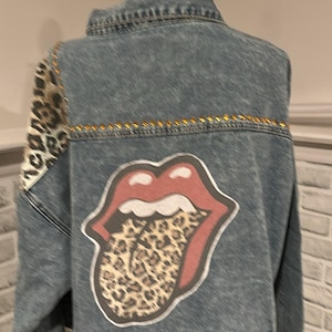May include: A denim jacket with a distressed look, featuring a leopard print sleeve and a large red lips graphic with a leopard print tongue. Gold studs line the back yoke.