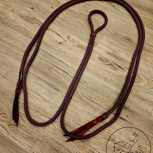 May include: A burgundy horse rein with a loop at one end and a leather accent at the other. The rope is thick and appears durable. The rein is lying on a wooden surface.