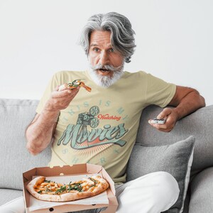 May include: A man wearing a pale yellow t-shirt with the text "This is my watching movies shirt" and a vintage film projector graphic. He is sitting on a grey sofa and holding a slice of pizza and a remote control.