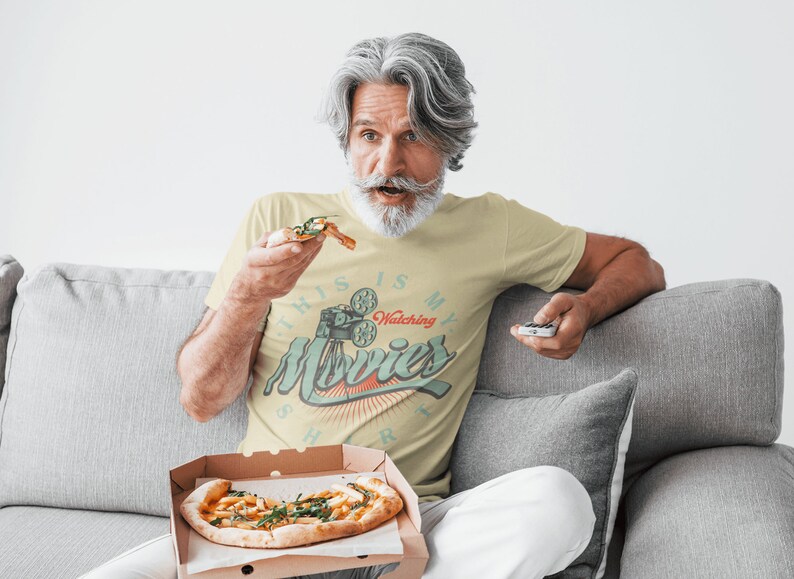 May include: A man wearing a pale yellow t-shirt with the text "This is my watching movies shirt" and a vintage film projector graphic. He is sitting on a grey sofa and holding a slice of pizza and a remote control.