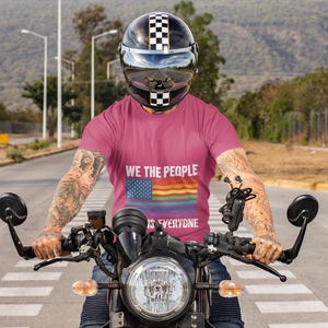 May include: A person on a motorcycle wearing a pink t-shirt with the text "WE THE PEOPLE is EVERYONE" and a rainbow flag design. The person is wearing a black and white checkered helmet. The motorcycle has a black headlight and handlebars.