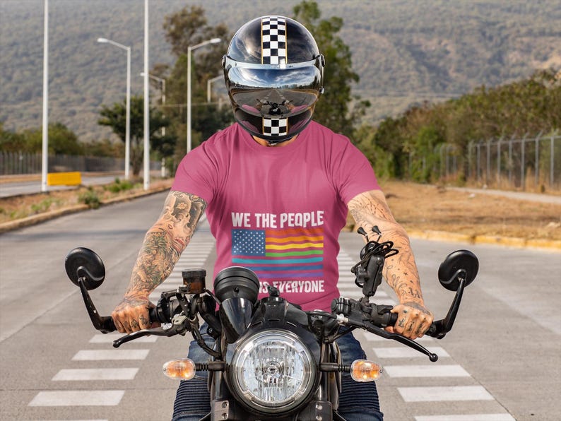 May include: A person on a motorcycle wearing a pink t-shirt with the text "WE THE PEOPLE is EVERYONE" and a rainbow flag design. The person is wearing a black and white checkered helmet. The motorcycle has a black headlight and handlebars.