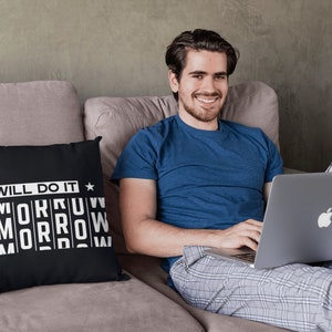 K&ouml;nnte beinhalten: Ein Mann sitzt auf einem Sofa mit einem Laptop auf dem Scho&szlig;. Er tr&auml;gt ein blaues T-Shirt und eine karierte Schlafhose. Ein schwarzes Kissen mit wei&szlig;em Text, der "Ich werde es morgen tun" lautet, liegt auf dem Sofa.
