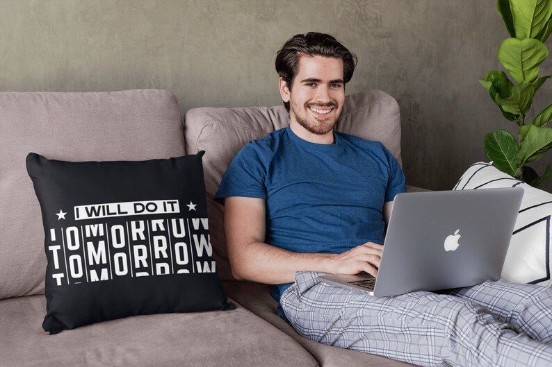 K&ouml;nnte beinhalten: Ein Mann sitzt auf einem Sofa mit einem Laptop auf dem Scho&szlig;. Er tr&auml;gt ein blaues T-Shirt und eine karierte Schlafhose. Ein schwarzes Kissen mit wei&szlig;em Text, der "Ich werde es morgen tun" lautet, liegt auf dem Sofa.