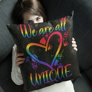Puede incluir: Una almohada negra con el texto "We are all UNIQUE" en colores arco&iacute;ris y un gr&aacute;fico de coraz&oacute;n arco&iacute;ris.