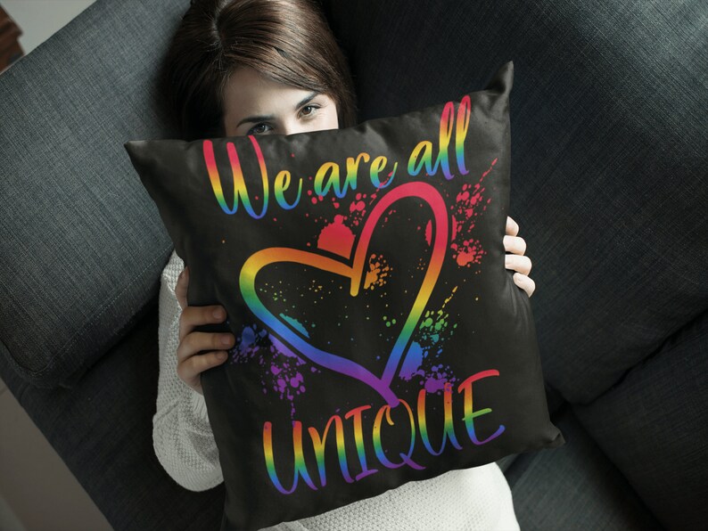 Puede incluir: Una almohada negra con el texto "We are all UNIQUE" en colores arco&iacute;ris y un gr&aacute;fico de coraz&oacute;n arco&iacute;ris.