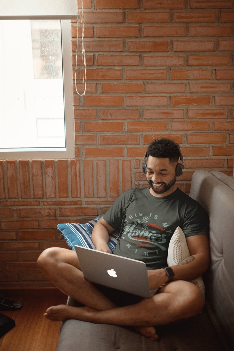 May include: A person wearing a dark green t-shirt with the text "This is my movies shirt" and a film reel graphic. The person is sitting on a sofa with a laptop on their lap and headphones on their head.