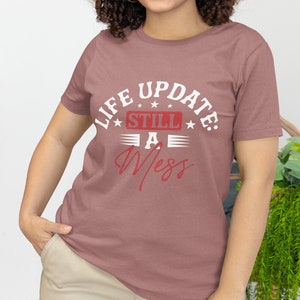 以下が含まれることがあります： マーブ色のTシャツに、「Life Update: Still A Mess」という文字が白と赤で書かれています。文字の周りは星とストライプで囲まれています。