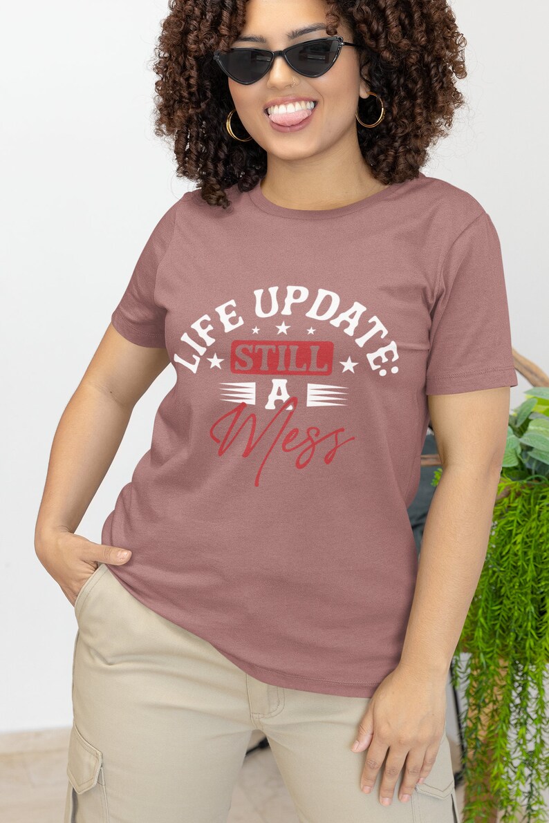 以下が含まれることがあります： マーブ色のTシャツに、「Life Update: Still A Mess」という文字が白と赤で書かれています。文字の周りは星とストライプで囲まれています。