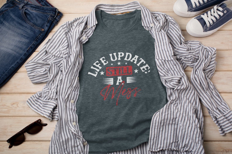 以下が含まれることがあります： 暗いグレーのTシャツに、「Life Update: Still a Mess」という白いグラフィックがプリントされています。グラフィックは赤い枠の中にあり、周りを白い星で囲まれています。Tシャツは、青と白のストライプのシャツ、青いジーンズ、青いスニーカーと一緒に、木製の表面に置かれています。