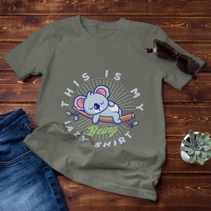 以下が含まれることがあります： 枝で眠る漫画のオウムのイラストが描かれた緑色のTシャツ。Tシャツには「This is my lazy being shirt」という文字が書かれています。