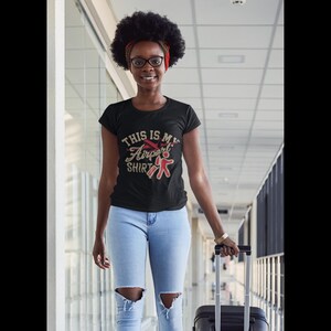 Puede incluir: Una mujer con una camiseta negra con el texto "This is my Airport Shirt" y un gr&aacute;fico de avi&oacute;n rojo. Lleva pantalones vaqueros azules y lleva una maleta negra.