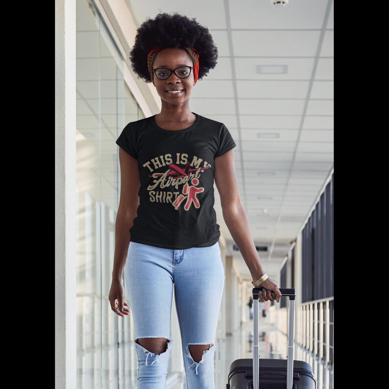 Puede incluir: Una mujer con una camiseta negra con el texto "This is my Airport Shirt" y un gr&aacute;fico de avi&oacute;n rojo. Lleva pantalones vaqueros azules y lleva una maleta negra.